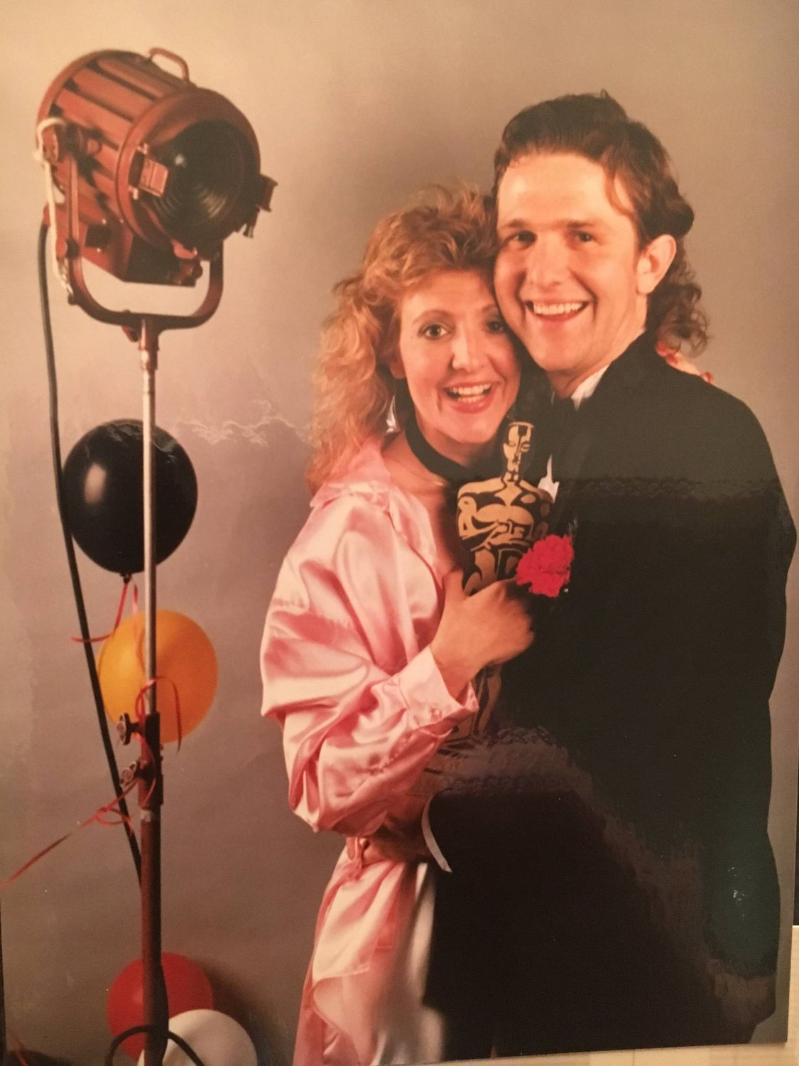 A couple in 1980s formal wear holding a golden statuette beside a film light and balloons