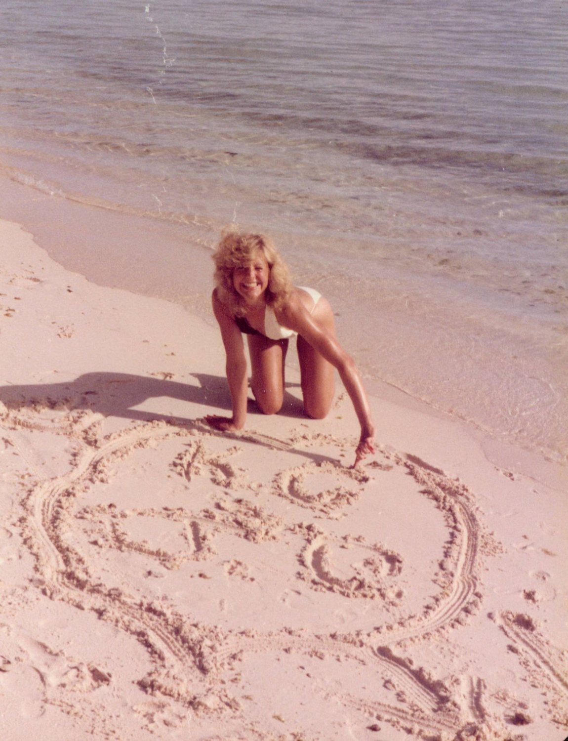 A smiling woman kneeling on a beach beside a drawing in the sand