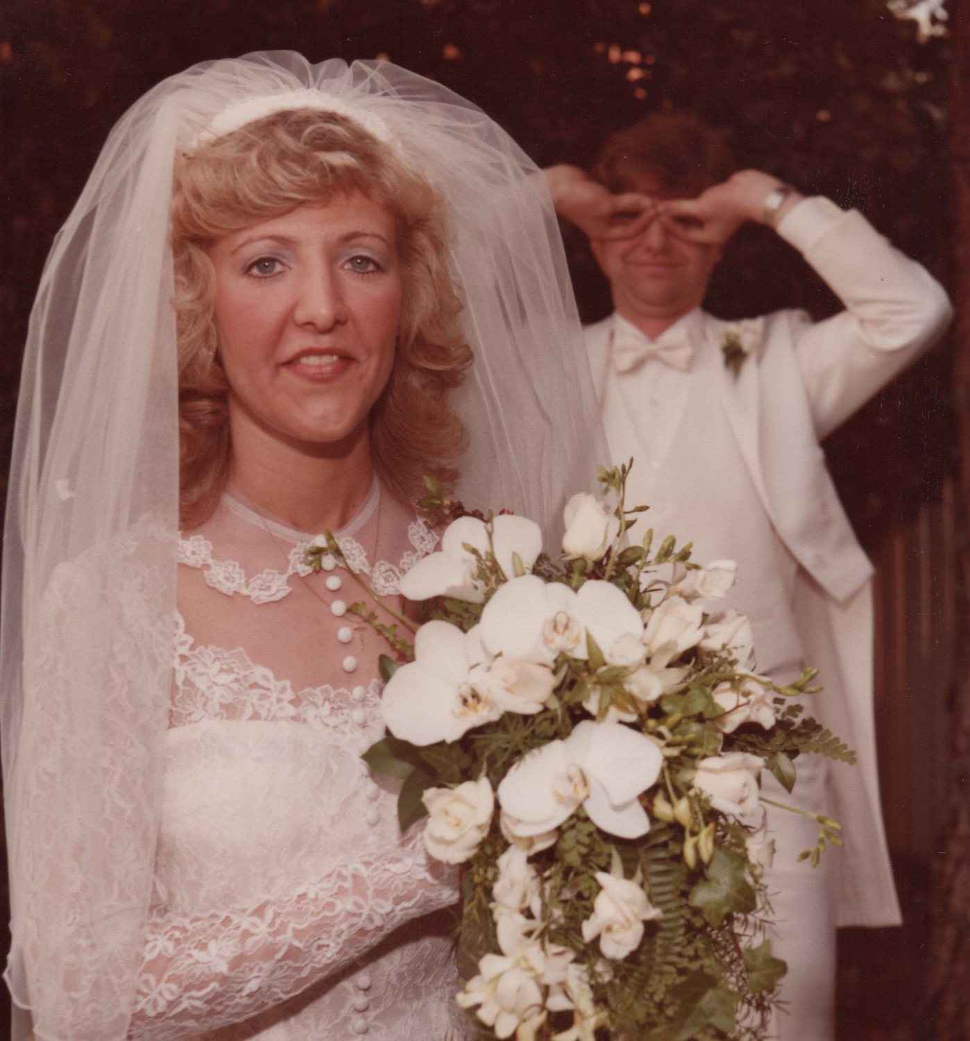 A bride with her bouquet in front, the groom behind her making binoculars with his hands