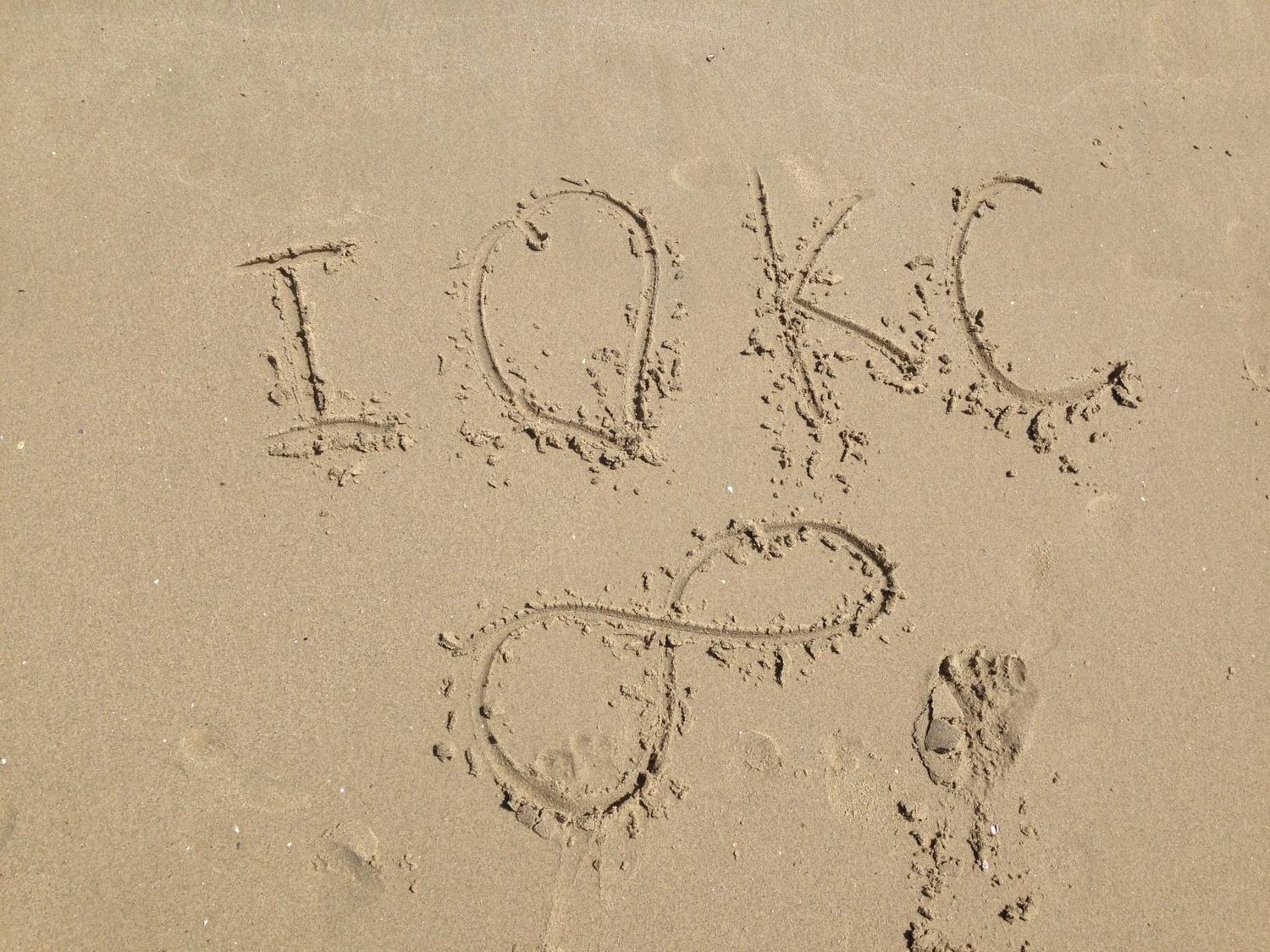 I, a heart, and KC written in the sand, with an infinity sign below
